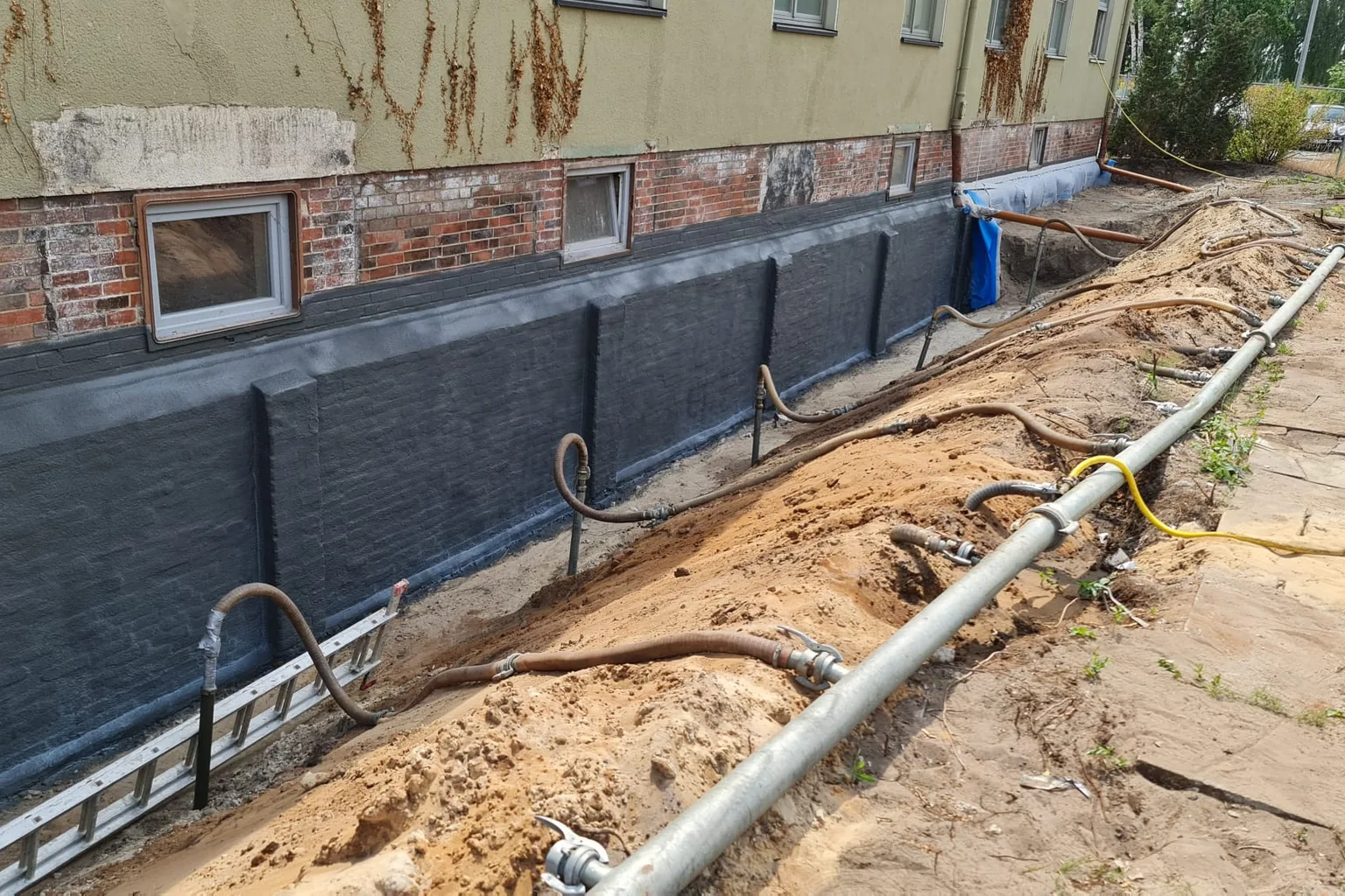 Zwei Handwerker dichten eine Betonkellerwand im Arbeitsgraben ab